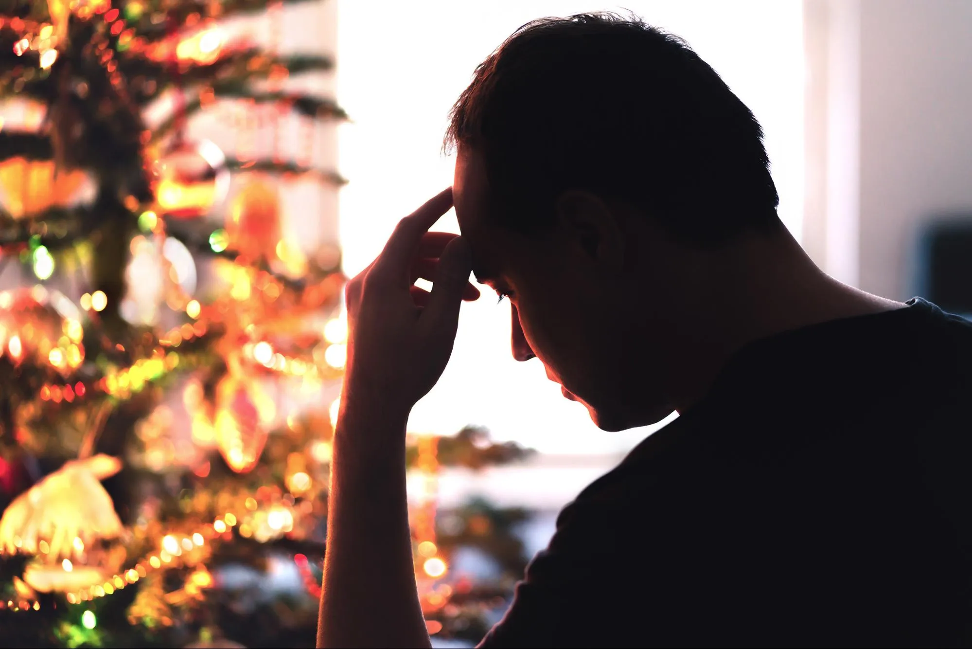 A silhouette of a person deep in thought, touching their forehead near a softly lit Christmas tree, creating a contemplative and warm atmosphere.